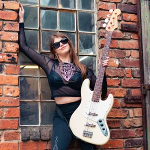 Keira Kenworthy with white Fender Jazz Bass and Rotosound Swing Bass 66 strings. Female bassist wearing sunglasses